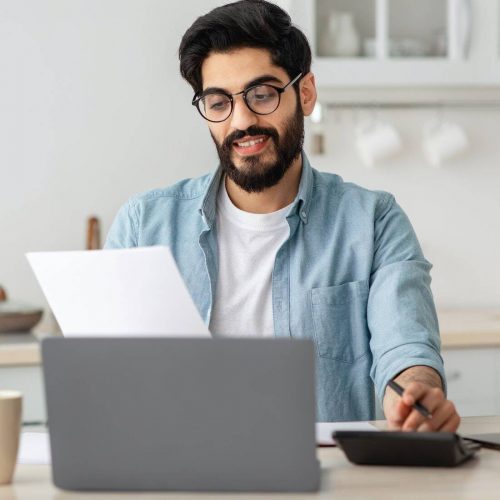 Portrait of happy arab guy reading insurance documents at home, sitting at table in kitchen and